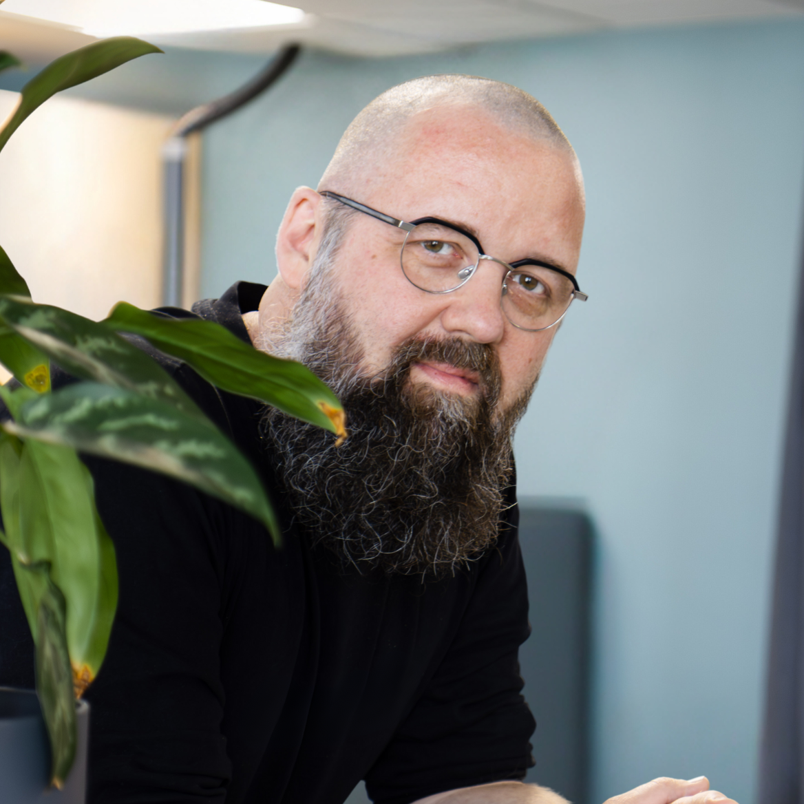 Mika Vaasala, Digitekoja oy perustaja, nojaa rennosti pöytään sisätiloissa. Hänellä on silmälasit ja tuuhea parta. Edustalla näkyy vihreä kasvi ja vaaleansininen seinä.