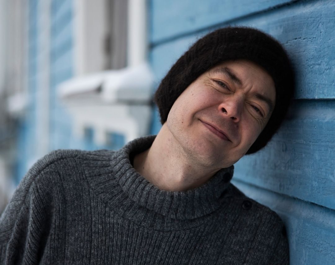 Vierakynäkirjoittaja Kyösti Iivonen katselee hymyillen kohti kameraa ja nojaa päällään puutalon siniseen seinään. 